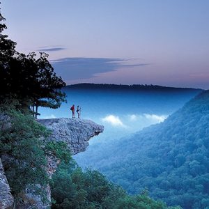 Two people standing on rock ledge looking over valley of tree covered hills