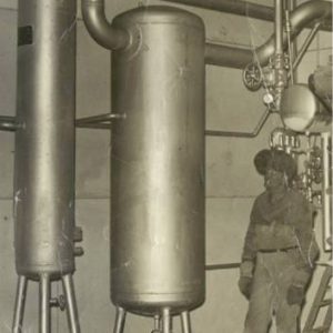 man in welding mask stands in front of large metal cylinders