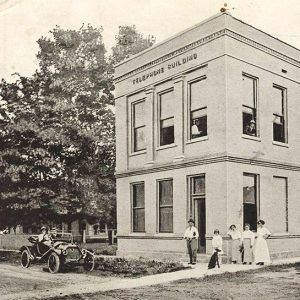 Two story white brick building with people standing on sidewalk and car parked in street