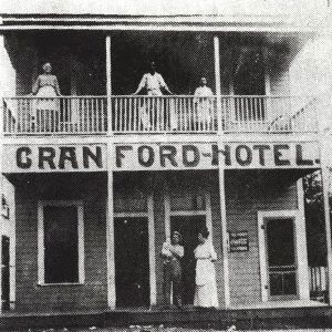 Two story wooden building with balcony with people standing out front and on the balcony