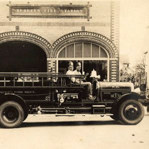 Two men sitting in fire truck in front of fire station