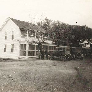 Multistory white wooden building with people on the sidewalk and cars parked alongside