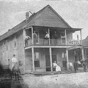 Two story wooden building with people on sidewalk and balcony along row of storefronts