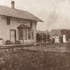 Single story wooden building beside railroad tracks "Cave Springs" with people standing on platform