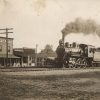 Train billowing smoke on tracks in front of row of wooden buildings