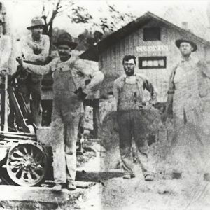 Six white men standing alongside machinery before single story wooden building