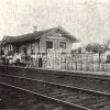 People and various barrels and other items around single story wooden building beside railroad tracks