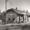 Four men standing before single story wooden building beside railroad tracks