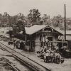 Wooden building beside railroad tracks with people and machinery around