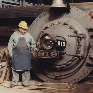 White man in hardhat and work clothes standing before locomotive components