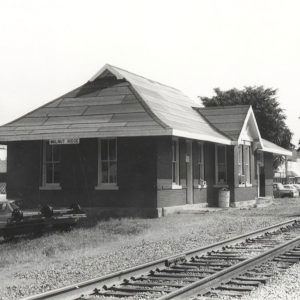 Single story brick building beside railroad tracks with a line of cars parked next to it