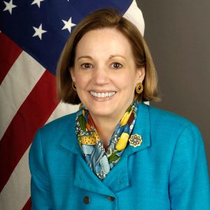 White woman in blue suit and scarf in front of American flag