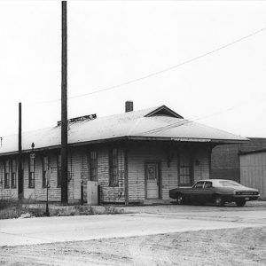 Single story wooden building next to railroad tracks with car parked at the end