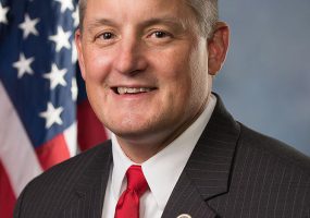 White man in suit and tie in front of American flag