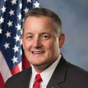 White man in suit and tie in front of American flag