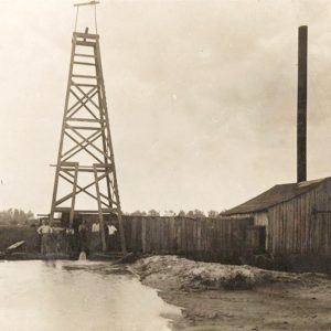 Man standing alongside latticed derrick behind pool of water with wooden fence and building