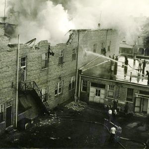 Partially collapsed building emitting smoke and men on roof of neighboring building spraying the former with hoses