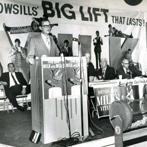 White man in suit speaking at lectern in front of promotional signs