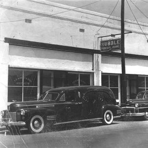 Multiple story brick building with hearse and another car parked out front