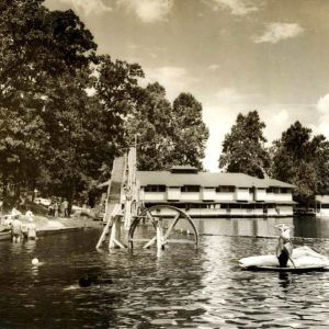 Tree-lined lake short with multistory building in distance and people playing in the water