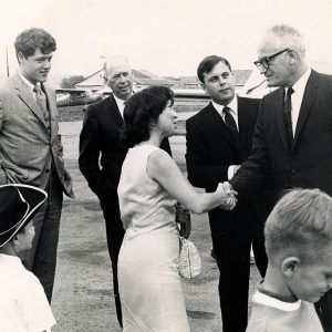 White woman at airport shaking hands with white man while other white men and white children watch