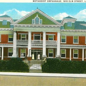 Two people standing and one person sitting on porch of multistory red brick building fronted with four columns