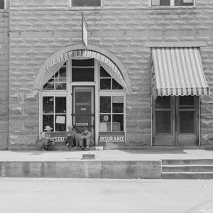 White men standing and sitting on sidewalk in front of multistory stone building