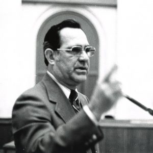 White man at microphone with finger raised