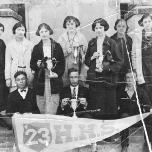 White students posing in front of school building with trophies and a banner.