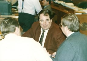 White man in glasses speaking with other white men in legislative chamber
