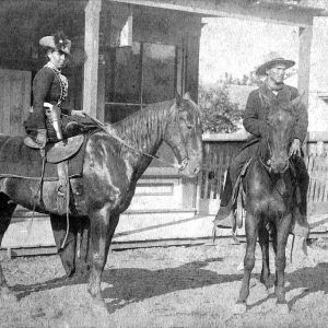 White woman wearing a hat and a gun holster and white man each on horses