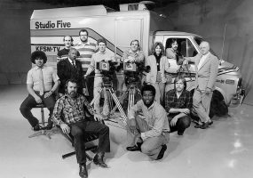 Large group of men and women standing before van with the words "Studio Five" on the side