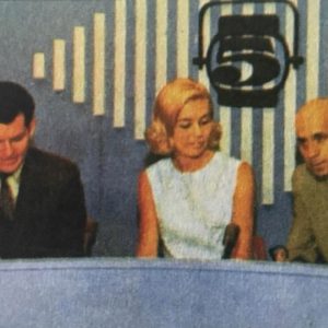 White woman in dress seated between two white men in television studio