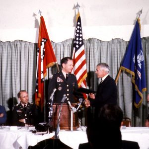 Two white men shaking hands at a lectern. Other white men seated at table