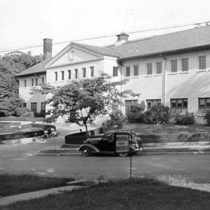 Multistory white building with car out front and man sitting on fence