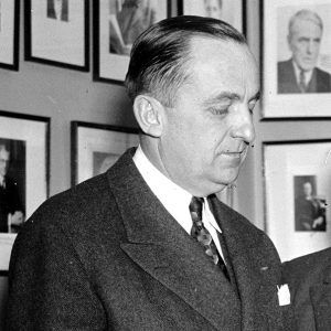 White man in suit and tie looking downward in front of wall with portraits