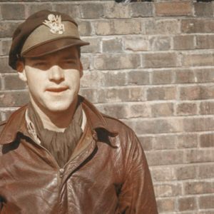 White man in military cap and leather jacket standing in front of brick wall