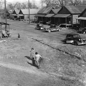 rows of wooden houses on road with a few people walking and sitting on a porch