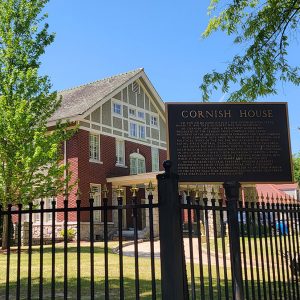 Multistory red brick building surrounded by black wrought iron fence with sign saying "Cornish House"