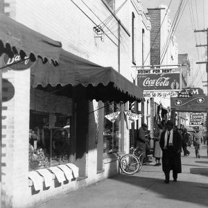 African American man in scarf walking down sidewalk next to buildings with signs for rooms for rent