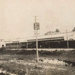 building with stacked lumber and train cars in front and water tower behind it