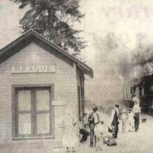 Small wooden building with sign saying "Prague" and several people waiting for arriving train