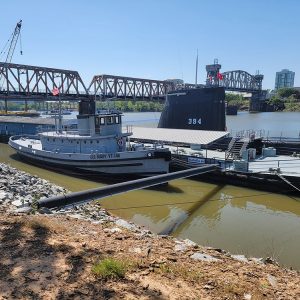 Tugboat and submarine docked on river with bridge in background