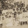 Large group of white children and adults beside wooden building