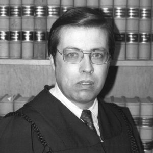 White man in glasses and judges robe standing in front of books