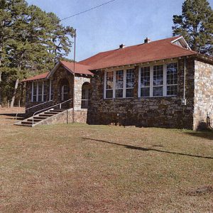 one-story rock school building with windows in front and red shingle roof and steps leading to front door