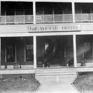 Multistory wooden building "Lafayette Hotel" with rocking chairs and people standing on lower porch