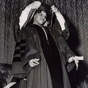 Young black woman in graduation robe while partially hidden white man puts academic hood around her neck