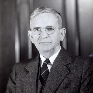 White man in suit and tie and glasses sitting at desk