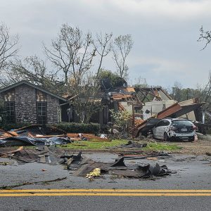 Home with roof torn off and some walls collapsed surrounded by debris and bare trees